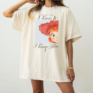 May include: An oversized cream-colored t-shirt featuring a graphic print of a cartoon character with red hair and a red hat. The text "EST 2026 I Knew It" appears above the graphic, and the text "I Knew You" is printed below the illustration.
