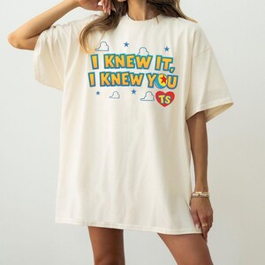 May include: An off-white short-sleeve t-shirt featuring the text I KNEW IT, I KNEW YOU in blue and yellow letters. The design includes small blue stars, white cloud outlines, and a red heart containing the letters TS, with a colorful ball graphic.