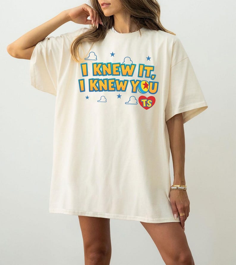 May include: An off-white short-sleeve t-shirt featuring the text I KNEW IT, I KNEW YOU in blue and yellow letters. The design includes small blue stars, white cloud outlines, and a red heart containing the letters TS, with a colorful ball graphic.
