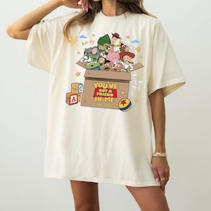 K&ouml;nnte beinhalten: Ein cremefarbenes Kurzarm-T-Shirt mit einer Grafik von Toy Story-Figuren, die aus einem Pappkarton kommen. Der Karton zeigt den Text "YOU'VE GOT A FRIEND IN ME" und "SINCE 1995", wobei "Andy" &uuml;ber den Figuren steht und Spielzeugw&uuml;rfel daneben liegen.