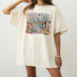 May include: An oversized cream-colored t-shirt featuring a colorful graphic print of various animated characters including Dumbo, Ariel, Cinderella, Mickey Mouse, Minnie Mouse, and Pluto, with the text Magic Kingdom printed below the characters.