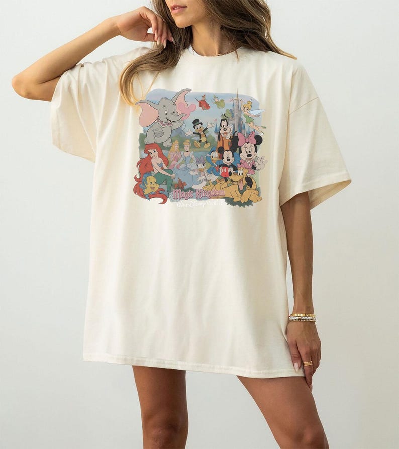 May include: An oversized cream-colored t-shirt featuring a colorful graphic print of various animated characters including Dumbo, Ariel, Cinderella, Mickey Mouse, Minnie Mouse, and Pluto, with the text Magic Kingdom printed below the characters.