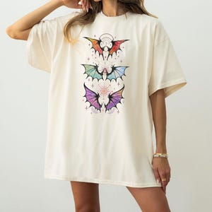 May include: Cream-colored oversized t-shirt featuring a colorful bat wing design. The graphic includes three bat wing illustrations in shades of red, green, and purple, with celestial accents. The shirt has short sleeves and a relaxed fit.