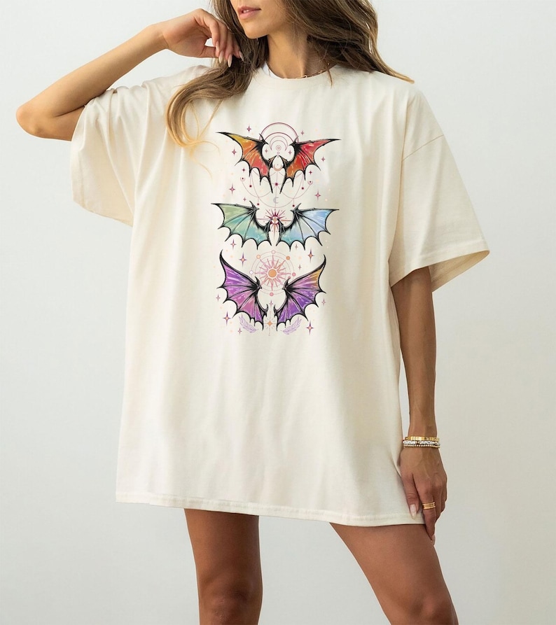 May include: Cream-colored oversized t-shirt featuring a colorful bat wing design. The graphic includes three bat wing illustrations in shades of red, green, and purple, with celestial accents. The shirt has short sleeves and a relaxed fit.