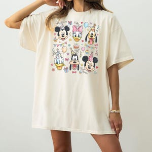 May include: Cream-colored t-shirt featuring a colorful graphic of Disney characters, including Mickey Mouse, Minnie Mouse, Donald Duck, Daisy Duck, Goofy, and Pluto, all wearing party hats. The design includes cupcakes, balloons, and confetti.