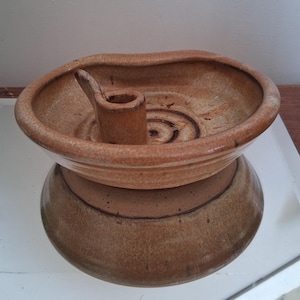 May include: A tan and brown ceramic candle holder with a wide, shallow bowl-shaped top and a central cylindrical socket. The interior base of the bowl features a spiral pattern, and the exterior has a smooth, rounded base with a distinct color transition.