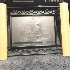 May include: A black metal photo frame with a rectangular aperture, supported by two light-coloured wooden posts. The frame has a wavy design. The frame is set against a dark background.