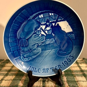 Könnte beinhalten: Ein blau-weißer Zierteller mit einer Szene mit Figuren und einer Katze. Der Teller hat einen Rand und den Text "JULE AFTEN 1963". Der Teller wird auf einem schwarzen Ständer präsentiert.