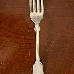 May include: A vintage silver fork with four tines and a decorative handle. The fork is made of a shiny metal and has a classic design. The handle has a rounded shape with a decorative element. The fork is likely used for eating.