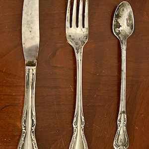 Puede incluir: Un juego de cubiertos antiguos de plata que incluye un cuchillo, un tenedor y una cuchara. Los utensilios presentan detalles ornamentales en el mango. El cuchillo está a la izquierda, el tenedor en el centro y la cuchara a la derecha.