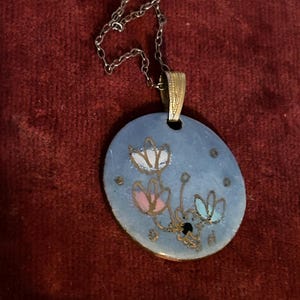 May include: A silver-toned necklace with a blue oval pendant. The pendant features a floral design in white, pink, and blue, with gold accents. The necklace is displayed on a red background.