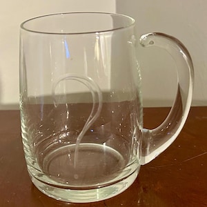 May include: A clear glass beer mug with a curved handle. A question mark is etched on the front of the mug. The mug sits on a wooden surface.