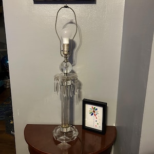 May include: A clear crystal table lamp with a bulb, a round crystal ball base, and hanging crystal accents. The lamp is on a dark brown half-moon table. A framed picture with the text "choose kind" is on the table.