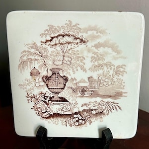 May include: Square ceramic tile with a brown pastoral scene featuring a large urn, trees, and architectural elements. The tile has a white background and a decorative border. The design is reminiscent of antique transferware.
