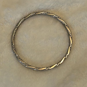 May include: A silver-colored bangle bracelet with a twisted rope design. The bracelet is circular and appears to be made of metal. The design features a series of interwoven lines, creating a textured surface. The bracelet is displayed on a light-colored, textured background.