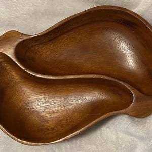 Puede incluir: Una fuente de madera para servir con una forma orgánica inusual. La fuente está dividida en dos secciones, cada una con un diseño curvo. La madera tiene un tono marrón cálido con vetas visibles, lo que sugiere un acabado natural. Adecuado para servir aperitivos o platos pequeños.