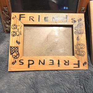 May include: A light brown wooden photo frame with the word "FRIENDS" written in black. The frame features small drawings of cartoon figures and objects. The frame has a rectangular aperture for a photograph.