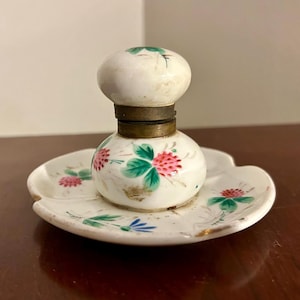 May include: Antique white porcelain inkwell with a matching saucer. The inkwell has a round body and a domed lid, connected by a brass collar. The inkwell and saucer are decorated with hand-painted pink flowers and green leaves.