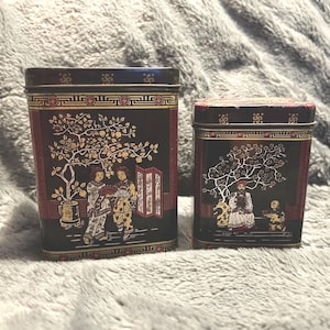 Pode incluir: Duas latas decorativas de metal preto com tampas. As latas apresentam uma borda vermelha e detalhes dourados. A lata maior mostra figuras sob uma árvore, a menor uma cena semelhante. As latas são retangulares.