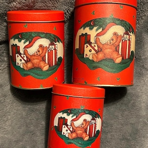 Peut inclure: Trois boîtes cylindriques rouges avec couvercles. Chaque boîte présente une illustration sur le thème de Noël d'un ours en peluche portant un chapeau de Père Noël, entouré de cadeaux emballés et de verdure. Les boîtes sont probablement destinées au stockage ou à la décoration.