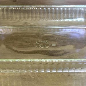 Puede incluir: Una fuente para hornear Pyrex rectangular transparente con lados y asas acanalados. El plato está hecho de vidrio y está diseñado para uso en horno. El plato está vacío y se encuentra sobre una superficie de madera.