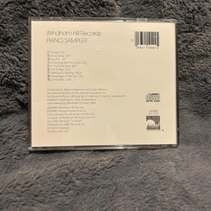 以下が含まれることがあります： 透明なプラスチックケースに入ったWindham Hill RecordsのピアノサンプラーCD。CDケースには、曲名とクレジットが記載された黒い文字の白いラベルが付いています。CDには「Compact Disc Digital Audio」と記載されています。