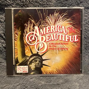 Puede incluir: Una caja de CD con el título "America The Beautiful" en letras rojas y blancas, con un fondo de fuegos artificiales y la Estatua de la Libertad. El logotipo de Reader's Digest Music es visible.