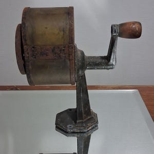 Puede incluir: Un sacapuntas manual vintage con cuerpo de metal oscuro y desgastado, que incluye un contenedor cilíndrico translúcido de tono ámbar. Posee una manivela lateral con un pomo de madera redondeado y una base octogonal resistente para su montaje.