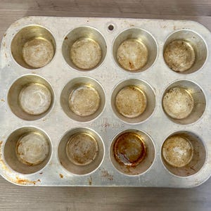 Könnte beinhalten: Eine gebrauchte, rechteckige Muffin-Form aus Aluminium mit zwölf kreisförmigen Formen. Die Form weist Gebrauchsspuren auf, mit Verfärbungen und Rückständen in den Mulden. Der Markenname ist auf der Seite sichtbar.