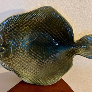 Może przedstawiać: Ceramiczny półmisek w kształcie ryby o teksturowanej powierzchni. Naczynie jest głównie ciemnozielone z niebieskimi i złotymi akcentami, posiada szczegółowe łuski i płetwy. Półmisek ma około 30 cm długości.