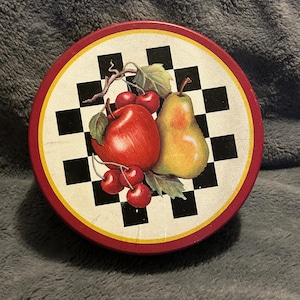 May include: Round decorative tin with a red border and a black and white checkered pattern. The center features a red apple, a yellow pear, and red cherries with green leaves. The tin is likely used for storage or display.