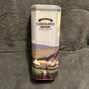 May include: An imported Carolans Finest Irish Cream tin with a landscape scene. The tin is a light blue color with gold trim. The landscape depicts a woman in a field, cottages, and rolling hills.