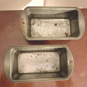 May include: Two vintage, rectangular loaf pans made of metal. The pans are a dull silver color with rounded edges and a small hole on each end. The interior of the pans shows signs of wear and use.