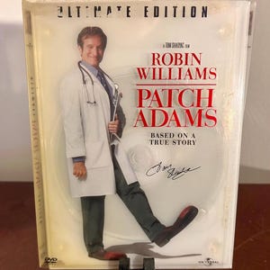 May include: DVD of the film "Patch Adams" starring Robin Williams. The cover features Williams in a doctor's coat, with the film's title in red and white. The DVD case is in a clear plastic sleeve.