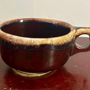 May include: A brown ceramic bowl with a handle. The bowl has a dark brown interior and a lighter brown rim. The handle is a rounded shape with a hole for gripping. The bowl sits on a gold-colored base.