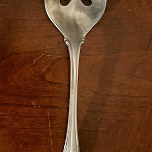 Puede incluir: Cuchara de servir plateada con un mango decorativo y un cuenco ovalado ranurado. La cuchara presenta un diseño floral en el mango y tres recortes decorativos en el cuenco. La cuchara está hecha de un metal brillante y reflectante.