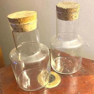 May include: Two clear glass bottles with cork stoppers. The bottles have a rounded base and a cylindrical neck. The cork stoppers are light brown. The bottles are on a dark wooden surface.