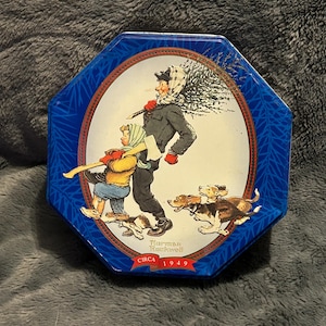 Könnte beinhalten: Achteckige blaue Dose mit einer Illustration von Norman Rockwell, die einen Mann und ein Kind mit einem Weihnachtsbaum und Hunden zeigt. Die Illustration ist in einem Oval mit rotem Rand gerahmt. Der Text "Circa 1949" ist sichtbar.