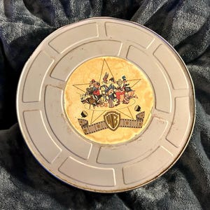 Puede incluir: Un contenedor de carrete de película plateado vintage con un diseño de personajes de dibujos animados de Warner Bros. de colores en el centro. El diseño presenta a Bugs Bunny, el Pato Lucas y otros personajes. El contenedor tiene forma circular.