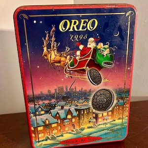 Op de afbeelding: Vintage Oreo koektrommel uit 1995. De trommel toont een feestelijke kerstscène met de kerstman in een slee getrokken door rendieren, vliegend over een besneeuwde stad. Het Oreo-logo is prominent aanwezig.