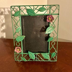 May include: Green metal picture frame with a lattice design, adorned with green leaves and pink and purple flowers. The frame has a dark interior and sits on a wooden surface.
