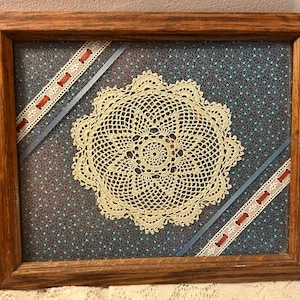 Può includere: Un'opera incorniciata con un centrino all'uncinetto color crema su tessuto floreale blu. La cornice in legno circonda il centrino. Nastri decorativi con dettagli di rocchetti rossi attraversano il tessuto.