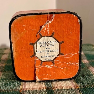 May include: An orange vintage box with black trim, featuring the text "QUELQUES FLEURS NATURELLE HOUBIGANT PARIS" in a white and black octagonal design. The box has visible wear and tear, with some of the orange paper peeling away.