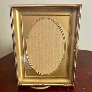 May include: A gold-colored picture frame with an oval opening. The frame has a rectangular shape with a gold border and a slightly distressed finish. The oval opening is filled with a tan-colored backing.