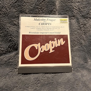 May include: A CD case featuring the album "Chopin" played by Malcolm Frager. The cover is a deep burgundy with the name "Chopin" in a stylized, off-white font. The CD case is in a clear plastic case.