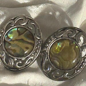 Puede incluir: Un par de pendientes ovalados con centros de concha de abulón iridiscente. Los pendientes tienen un diseño de filigrana plateado y ornamentado. La concha de abulón muestra una mezcla de tonos verdes, dorados y azules. Cada pendiente mide unos 2,5 cm.