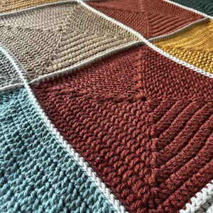 May include: A crocheted blanket with a patchwork design featuring squares in various colors, including mustard yellow, beige, red, teal, and white.