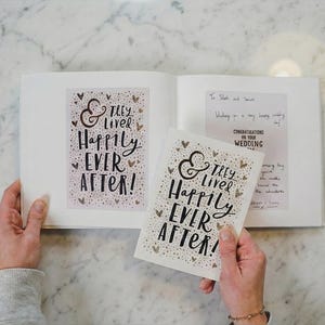 Könnte beinhalten: Ein offenes Buch und eine passende Karte mit dem schwarzen handgeschriebenen Text "& They Lived Happily Ever After!", umgeben von kleinen goldenen Herzillustrationen. Die Buchseite zeigt zudem eine handschriftliche Notiz mit dem Text "Congratulations on your wedding day."