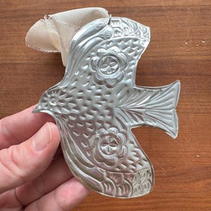 May include: A silver-colored metal bird ornament with embossed floral and feather details. The bird has a loop for hanging and is attached to a cream-colored fabric ribbon. The ornament is held in a hand against a wooden background.