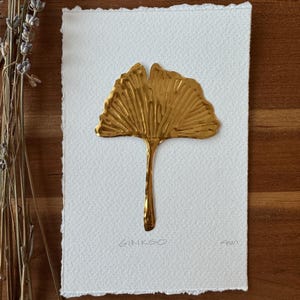以下が含まれることがあります： 金色のイチョウの葉の彫刻が、テクスチャ加工された白い紙の上にあります。「GINKGO」の文字が葉の下に書かれています。このアートワークは木製の背景にあり、左側には乾燥したラベンダーの小枝があります。